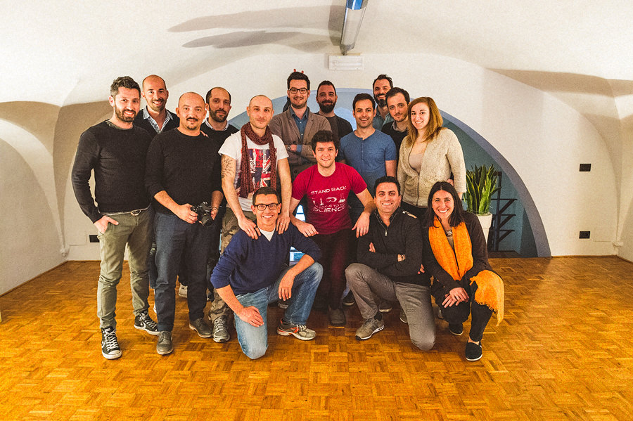 A group of 16 people poses for a photo in an indoor setting with arched ceilings. Fourteen people stand in the back, while two people kneel in front. They are all smiling and dressed casually.