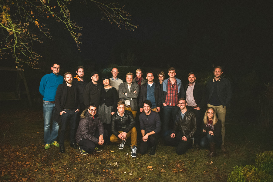 A group of 18 people posing together outdoors at night with some standing and some crouching, under tree branches with sparse leaves.