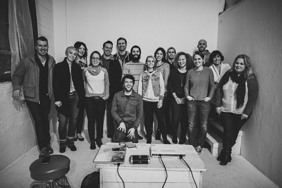 A group of 17 people, consisting of both men and women, standing and smiling indoors against a plain wall. One person is kneeling in the front, and there is a table with items on it in the foreground.