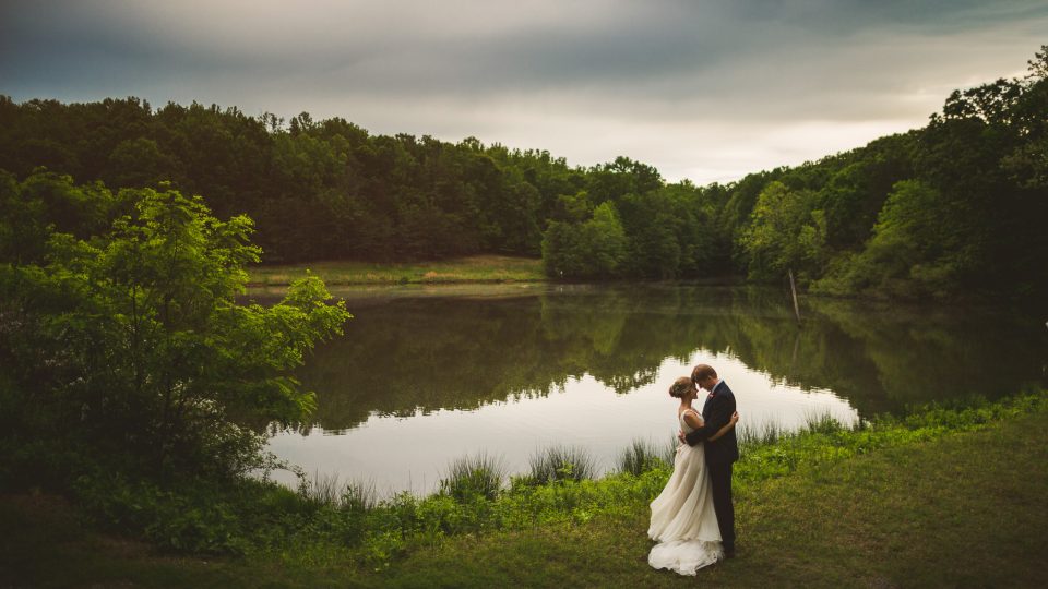 A bride and groom embrace near a tranquil lake surrounded by lush greenery under a cloudy sky, capturing the perfect wedding moment.