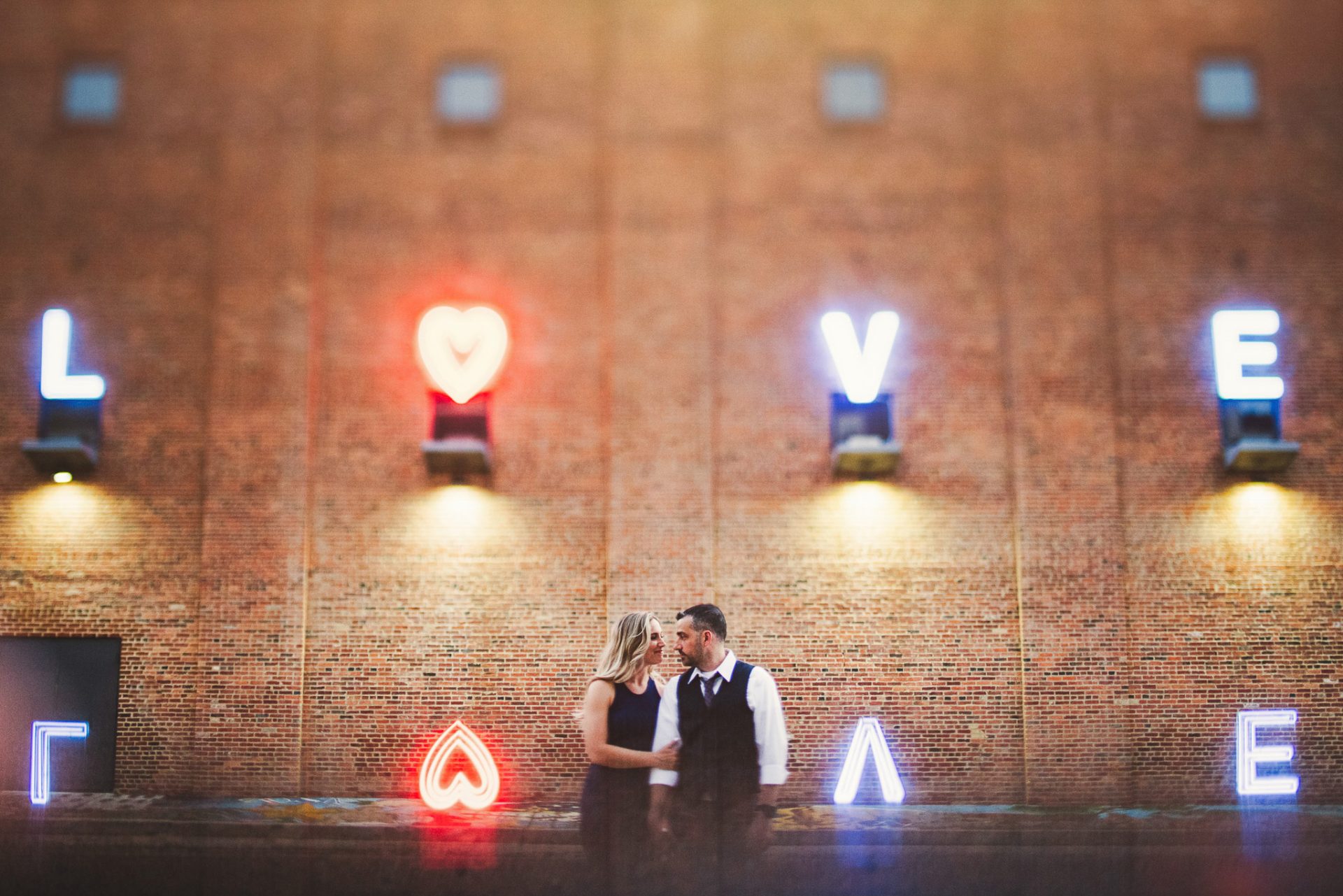 A couple stands together in front of a brick wall with neon lights spelling "LOVE," capturing a timeless wedding moment. This stunning photography would be perfect for any pricing guide, showcasing love's glow and passion.