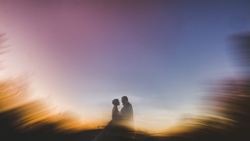 A silhouetted couple stands facing each other against a vibrant, blurred sunset sky in a perfect moment for timeless wedding photography.