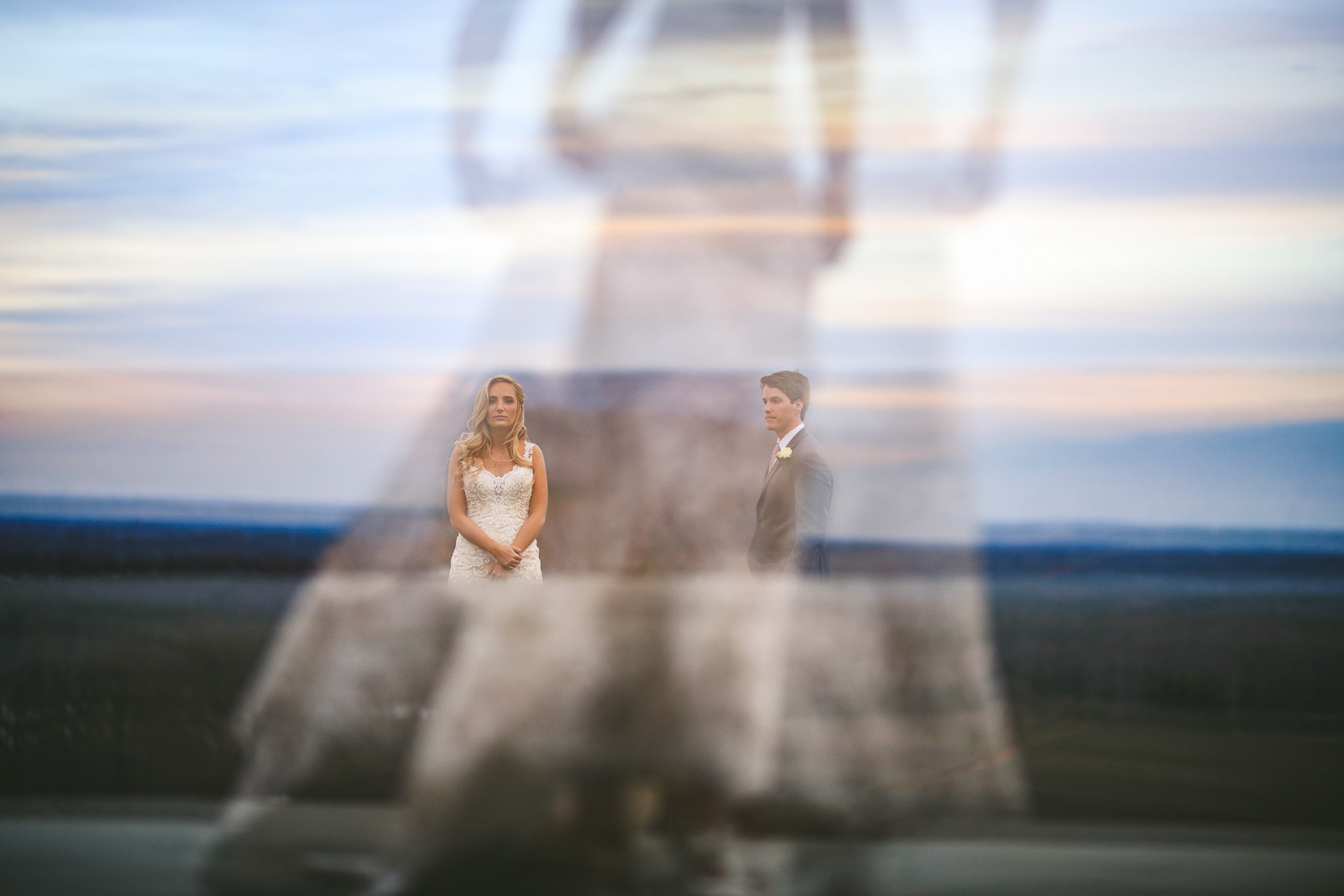 A bride and groom stand apart in an outdoor setting, with a soft, ghost-like overlay of the bride's dress in the foreground. The background is a blurred, expansive landscape under a cloudy sky—a dreamy moment captured perfectly by wedding photography. For more details, see our Pricing Guide.