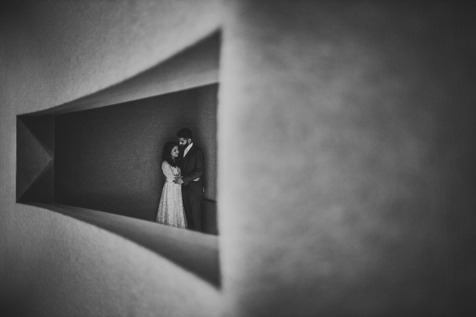 A couple stands closely together, seen through a triangular opening in a wall, with a minimalist and monochromatic aesthetic, evoking the timeless charm of wedding photography.