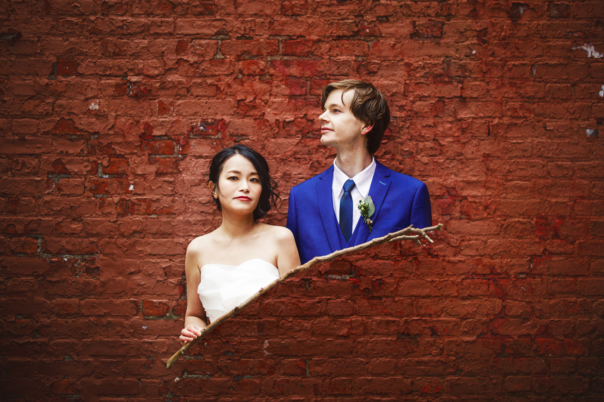 A bride and groom stand against a textured red brick wall with a gap, the groom in a blue suit and the bride in a white dress. Capture this beautiful wedding moment with our top-tier photography services.