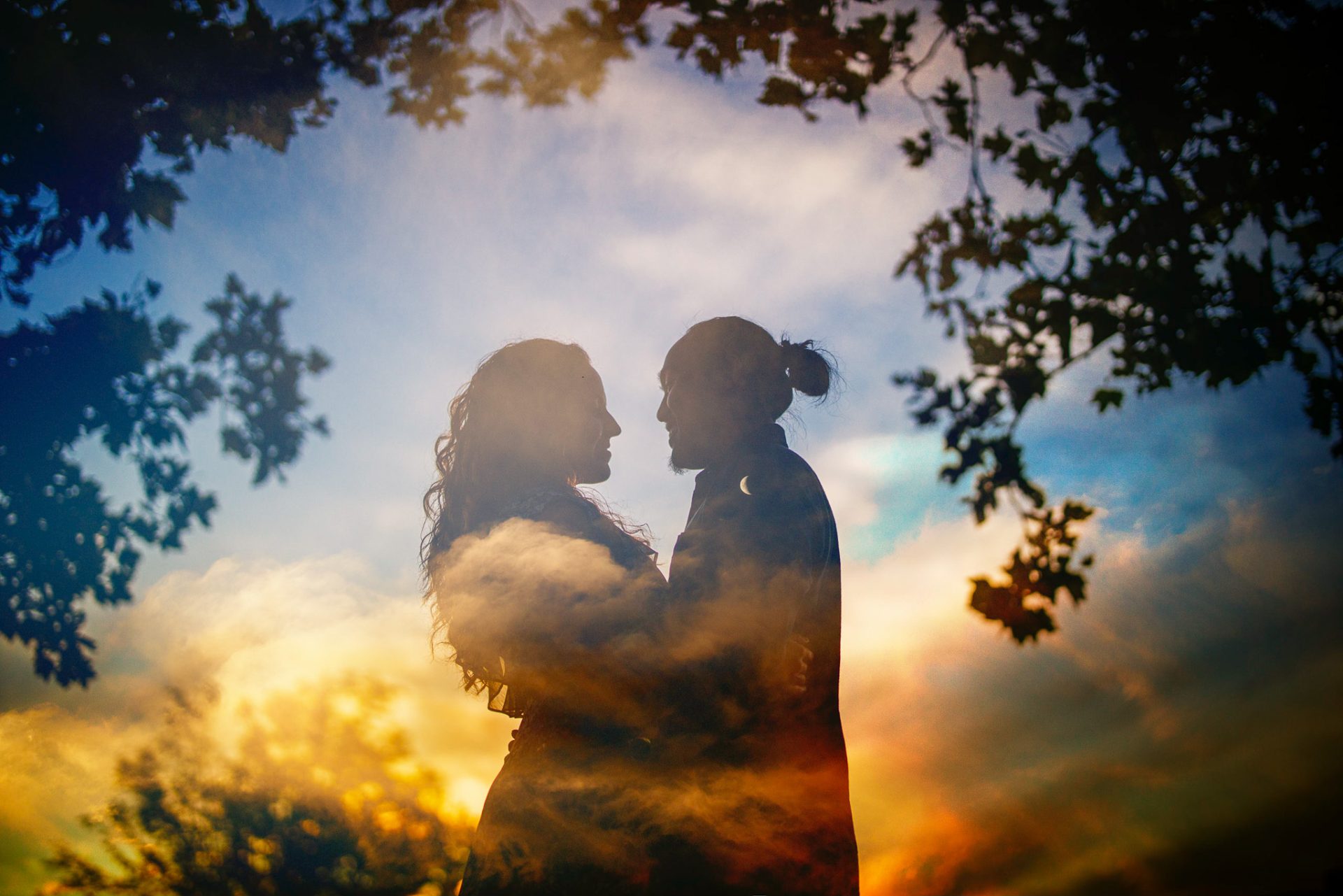 Silhouette of two people standing face to face surrounded by a colorful, cloudy sky and tree branches in the foreground—a perfect moment for wedding photography.