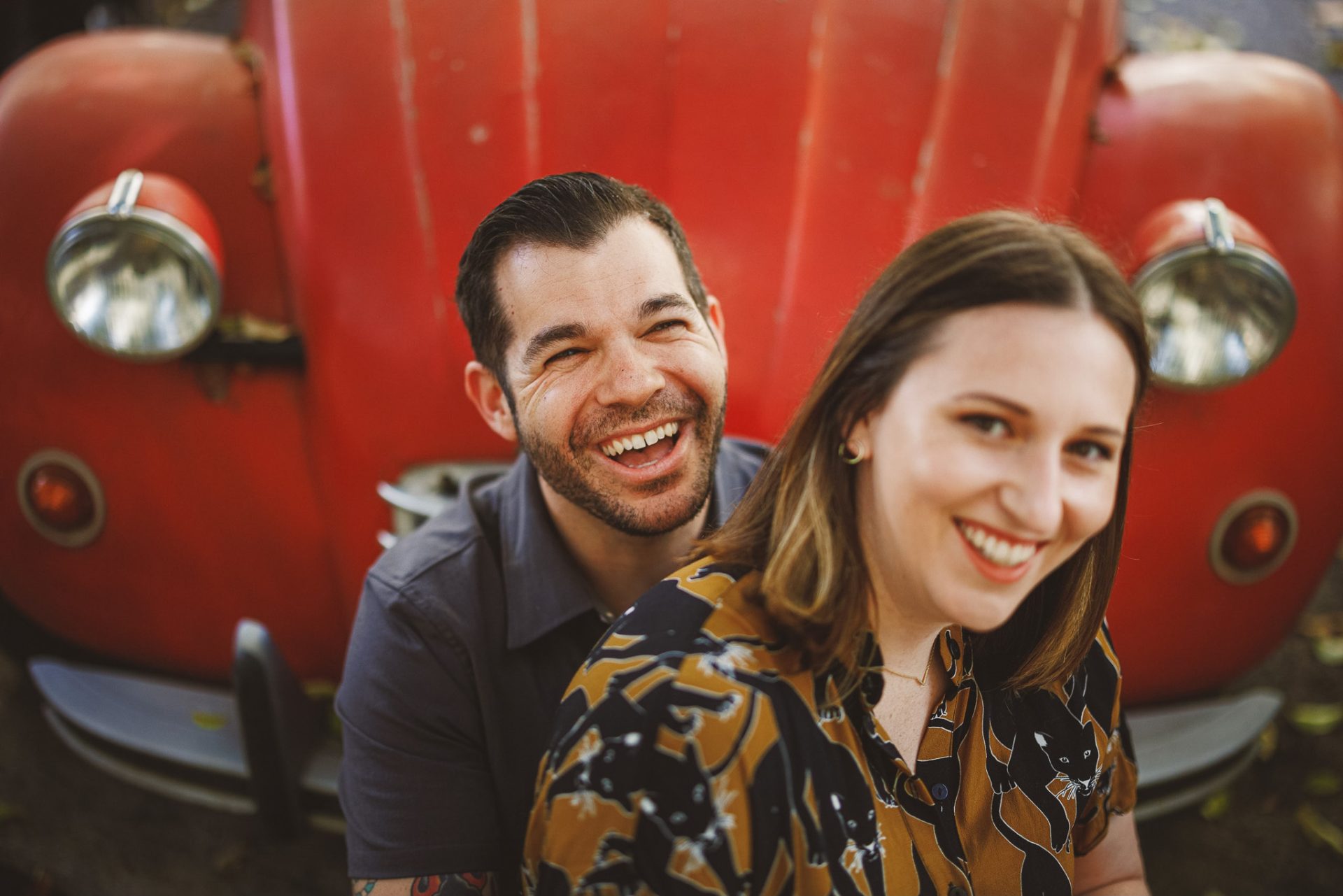 Two people sitting and smiling with a large red vintage car in the background, capturing a perfect moment that could grace the pages of any wedding photography portfolio.