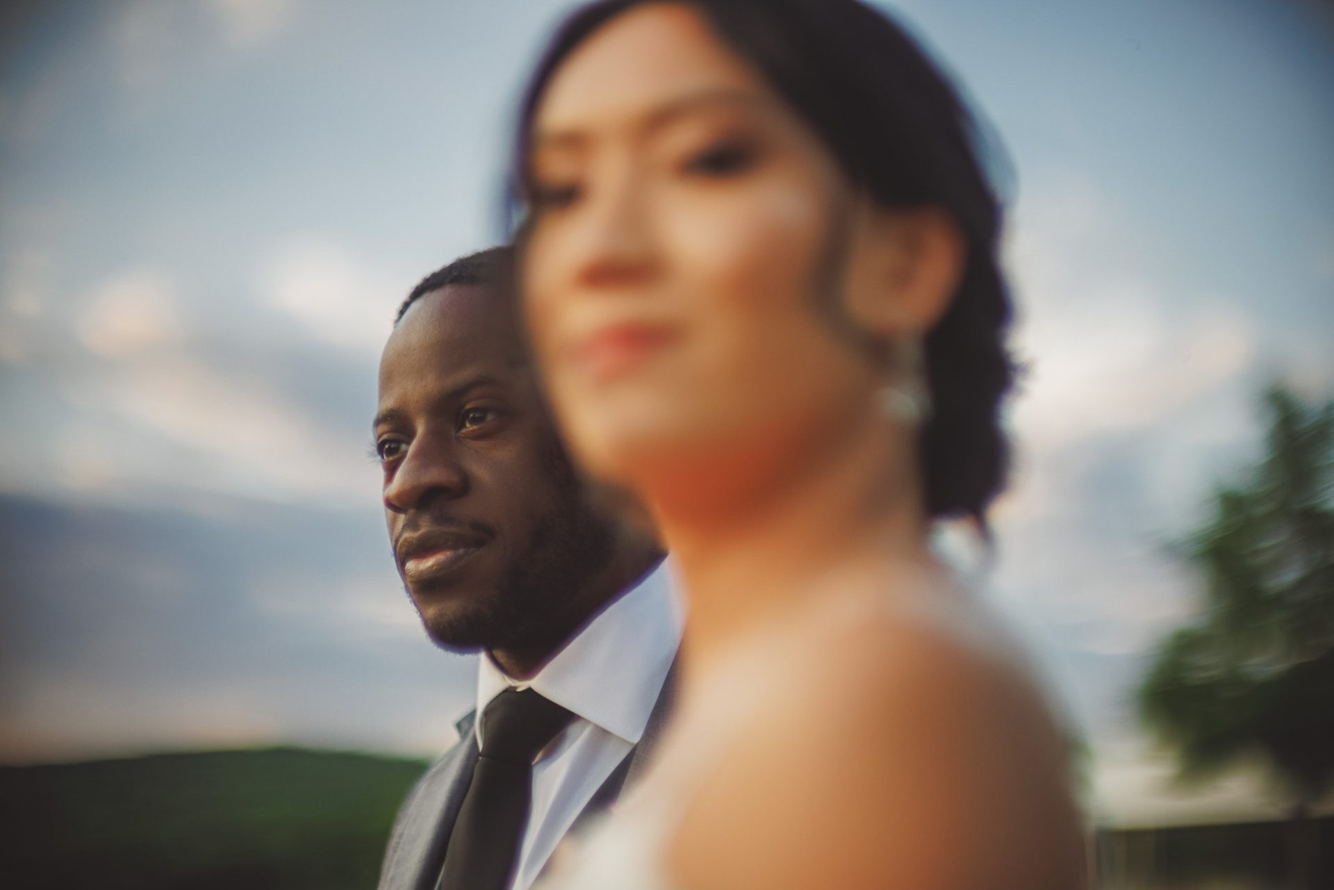 A man and woman stand outdoors, with the man in focus wearing a suit and the woman out of focus in the foreground. The background features a cloudy sky and blurred landscape, capturing a moment reminiscent of wedding photography.