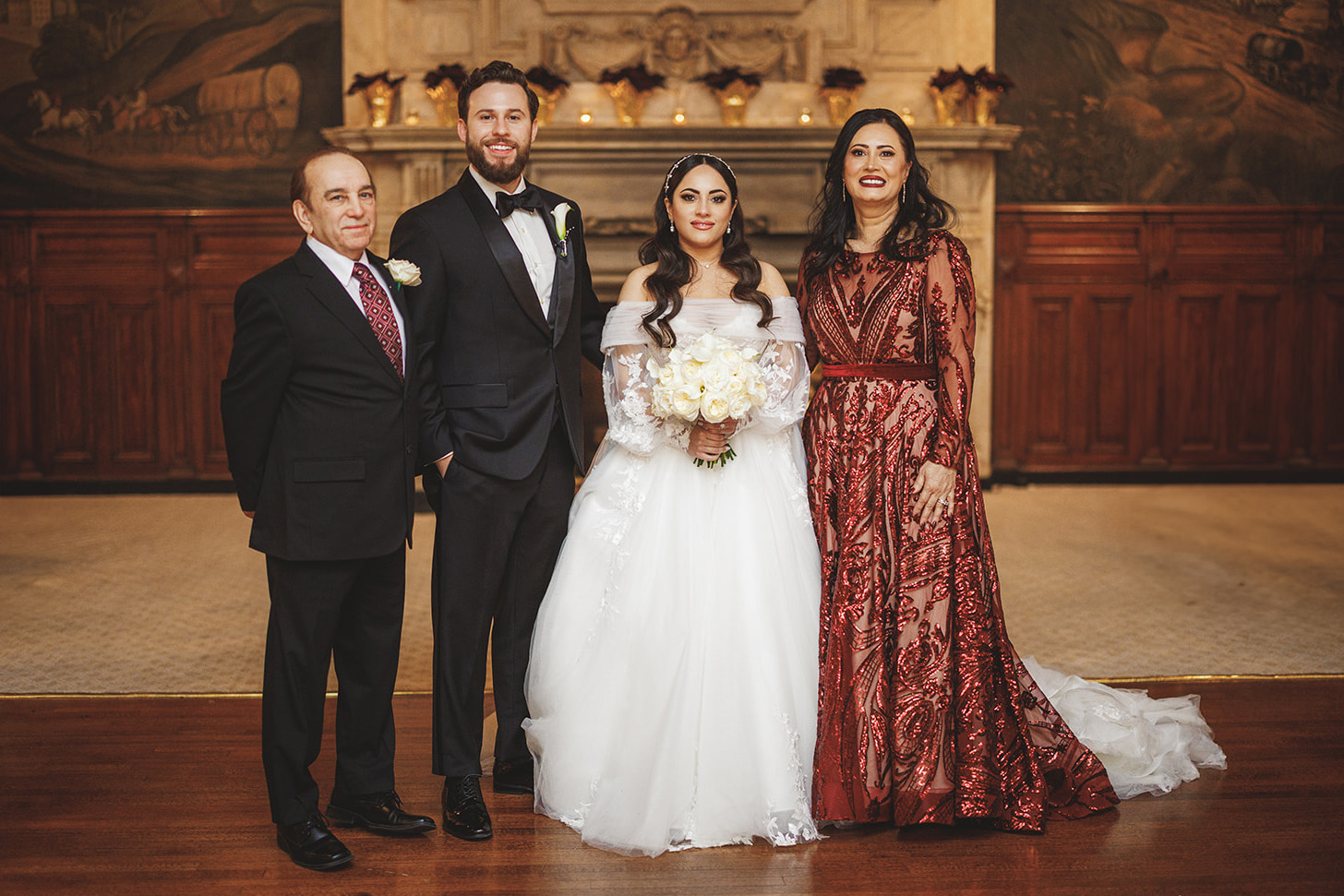 A bride and groom stand between two older adults in formal attire, posing in a grand room with a decorative fireplace in the background.