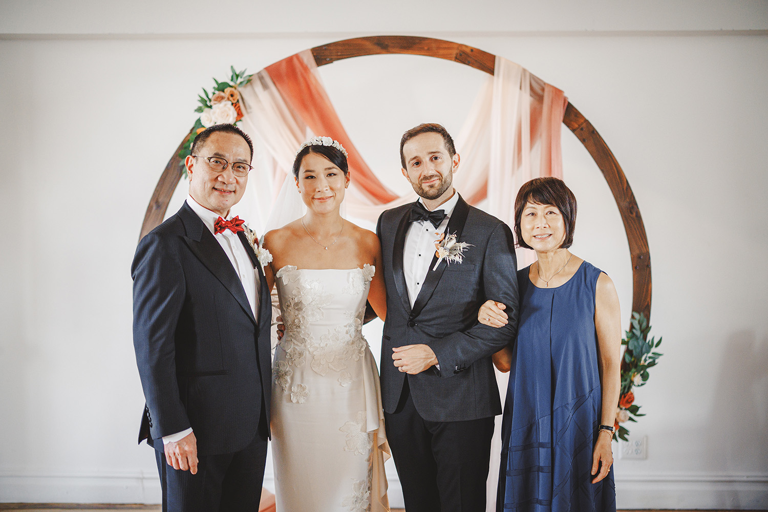 Four people stand in front of a wooden circular wedding arch with floral decorations. The bride and groom are in the center, flanked by an older man and woman. Everyone is dressed formally.