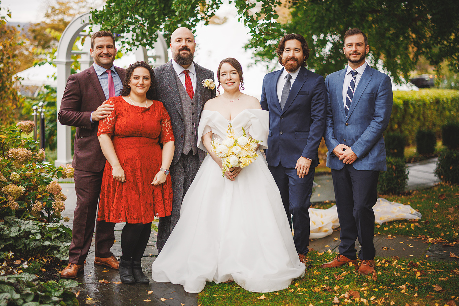 A bride and groom stand together with four guests, in an outdoor garden setting, all smiling for a group photo.