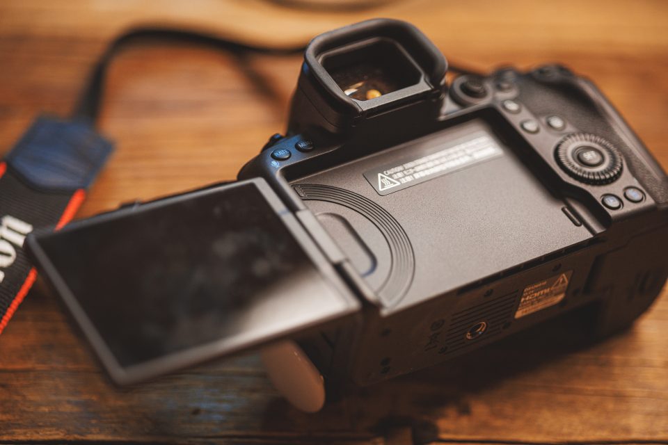 The Canon R5 Mark II, designed for real-world use, sits on a wooden surface with its rear viewfinder screen flipped out. The camera strap is partially visible, embodying a practical setup for any thorough review.
