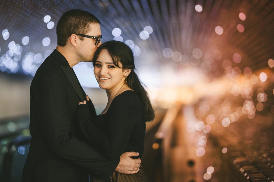 A man kisses a woman's forehead as they stand in an illuminated tunnel with bokeh lights in the background.