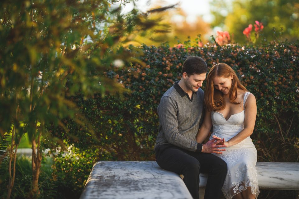 A couple sits on a stone bench in a garden, smiling and holding hands. The woman wears a white dress, and the man wears a grey sweater.