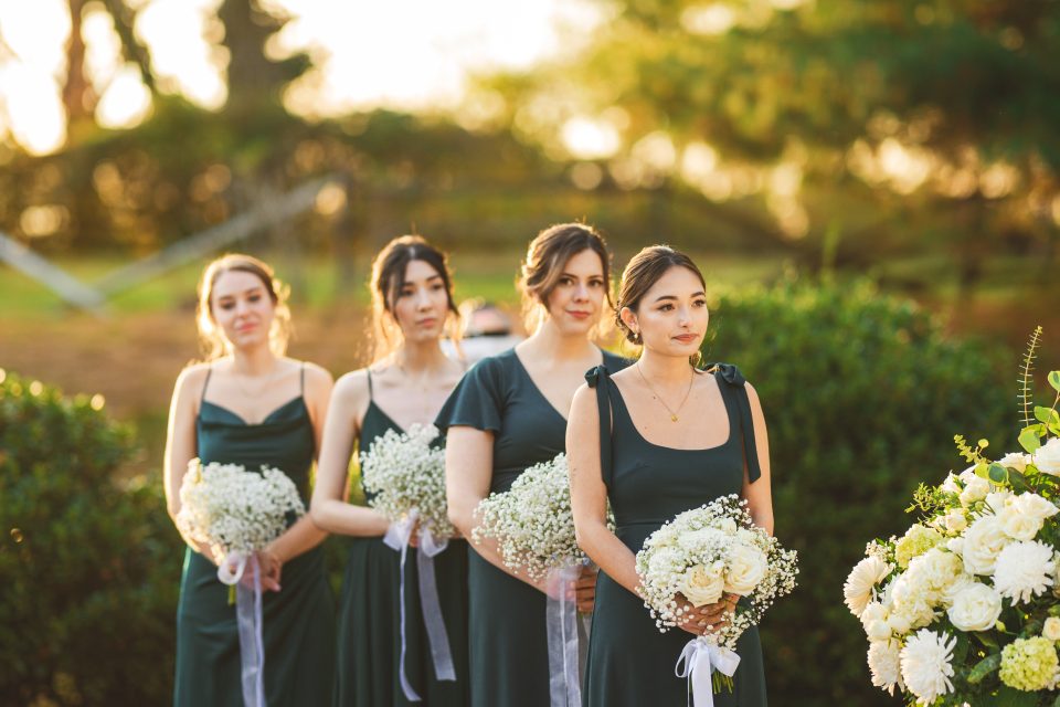 Four bridesmaids in dark green dresses holding white floral bouquets stand outdoors in a line.