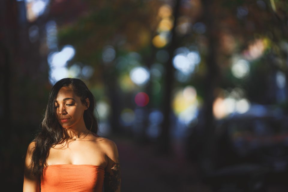 Woman in an orange top stands outdoors with eyes closed, surrounded by blurred trees and soft light.