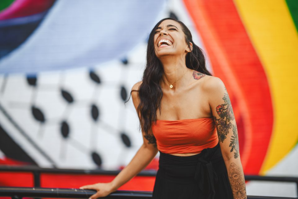 Person laughing with long dark hair, wearing an orange top and black pants, standing in front of a colorful mural.