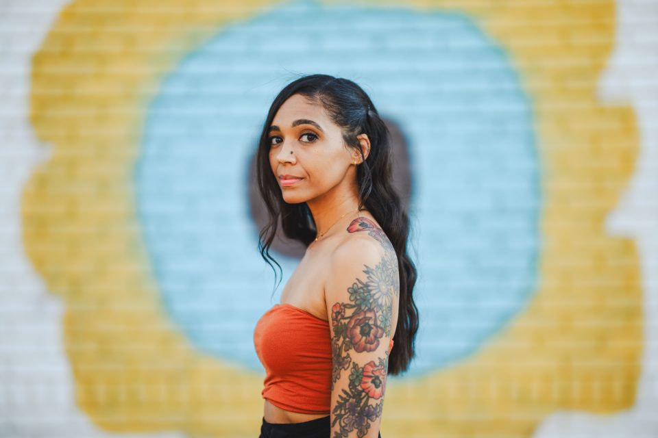 Woman with long hair and a floral tattoo on her arm stands in front of a colorful mural, wearing an orange top.