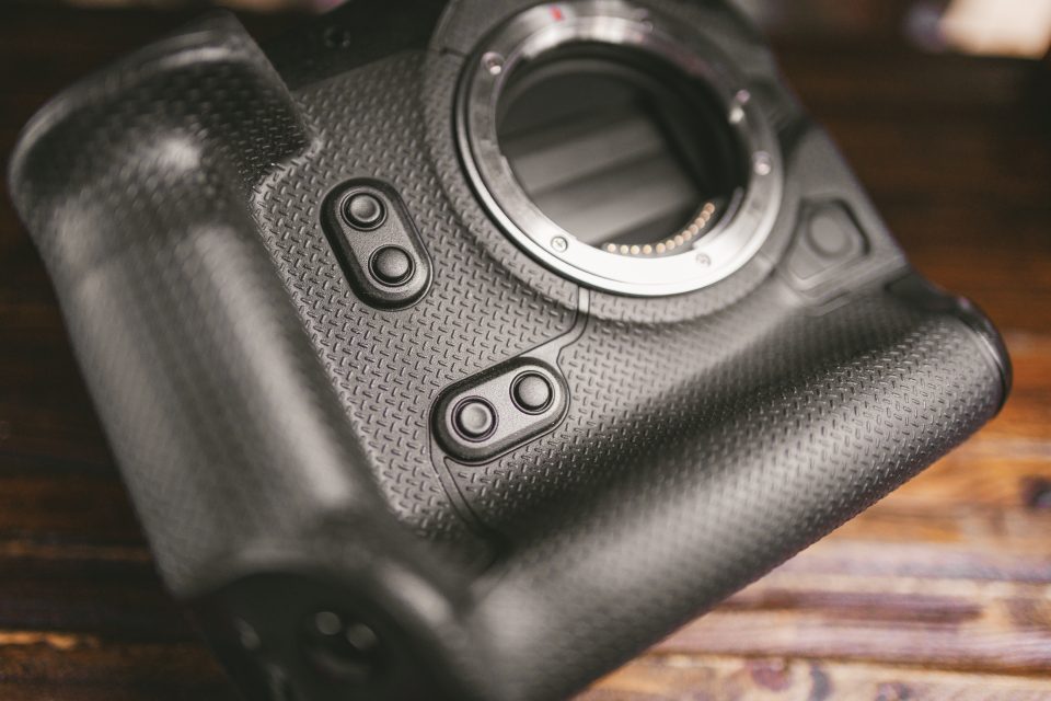 Close-up of a black digital camera body without a lens, showing textured grip, buttons, and part of the lens mount on a wooden surface.
