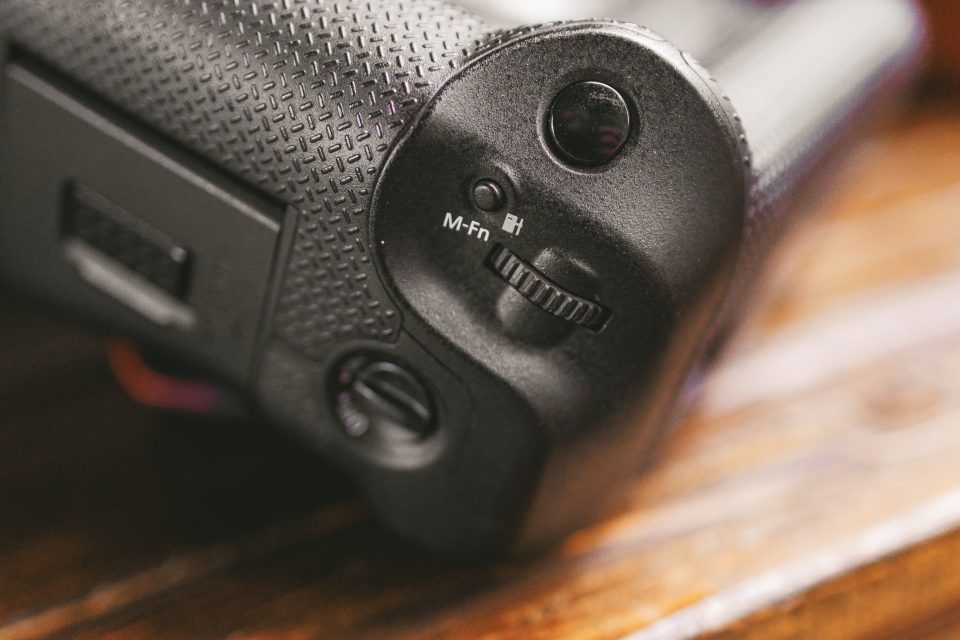Close-up of a textured camera grip showing a control dial, M-Fn button, and part of a switch, resting on a wooden surface.