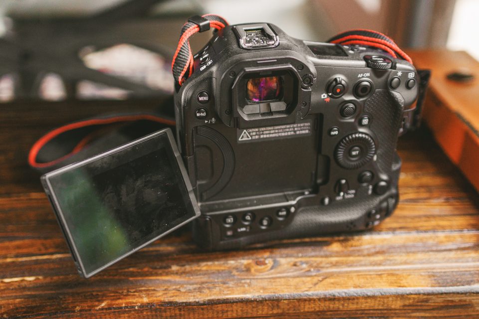 A digital camera with a flip-out LCD screen is placed on a wooden surface, showing the back view with various control buttons and a red-striped neck strap.