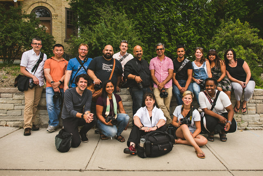 Group of 16 people, some standing and some seated on a stone ledge, posing outdoors with trees and a building in the background. Many are carrying cameras and bags.