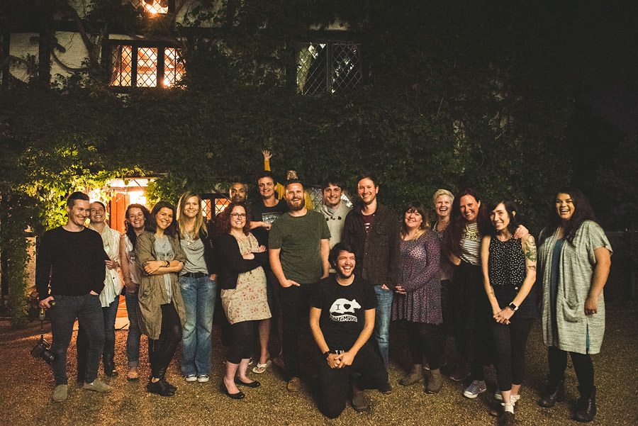 A group of 17 people gathers for a photo outside a building at night. Most are standing, one person sits in the front, and all are smiling. The building is covered in foliage and softly lit.