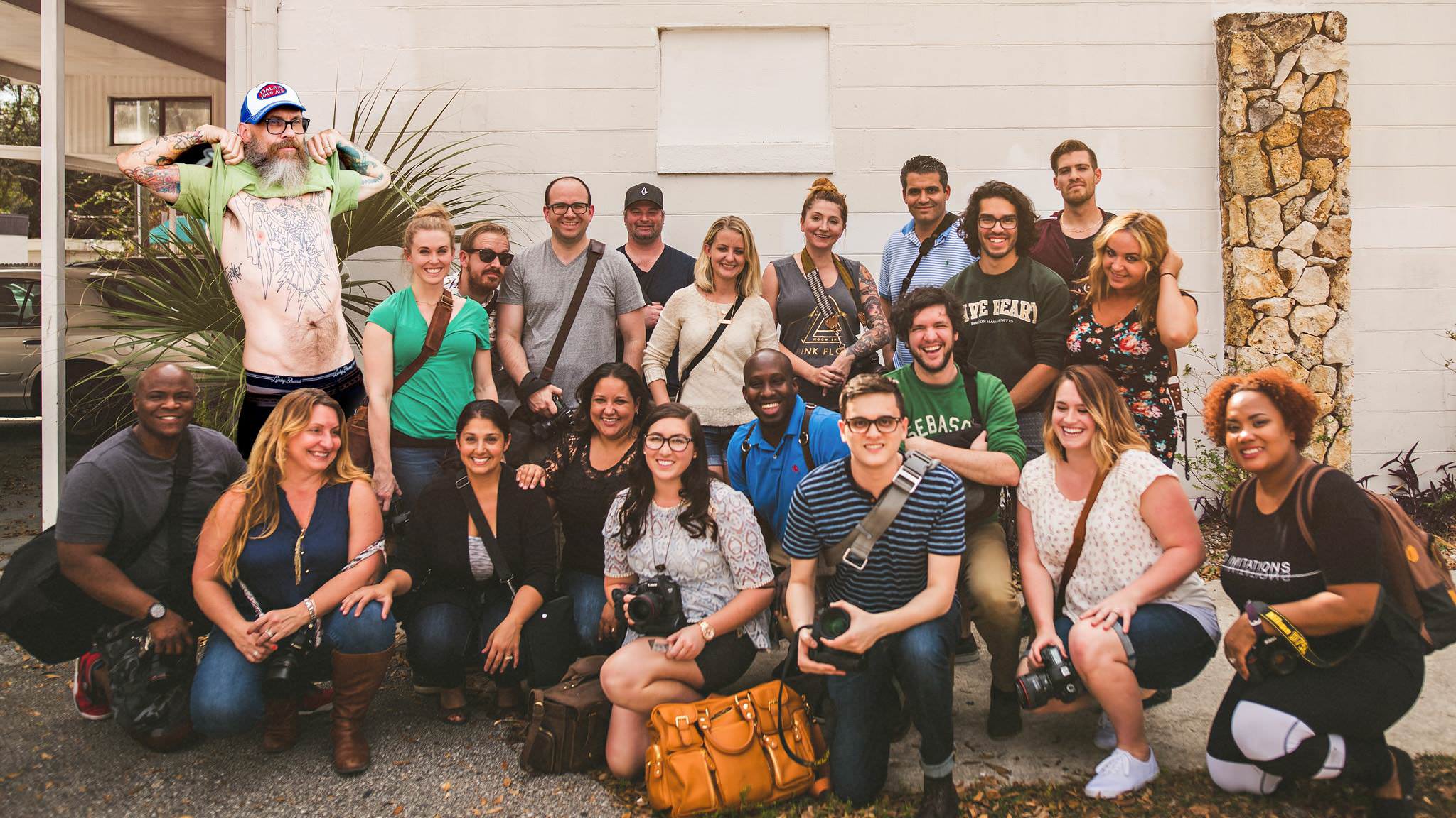 A group of 20 people posing for a photo outdoors. Some hold cameras, and one person on the left is wearing a hat and beard and is shirtless with visible tattoos. A plant and a stone wall are in the background.