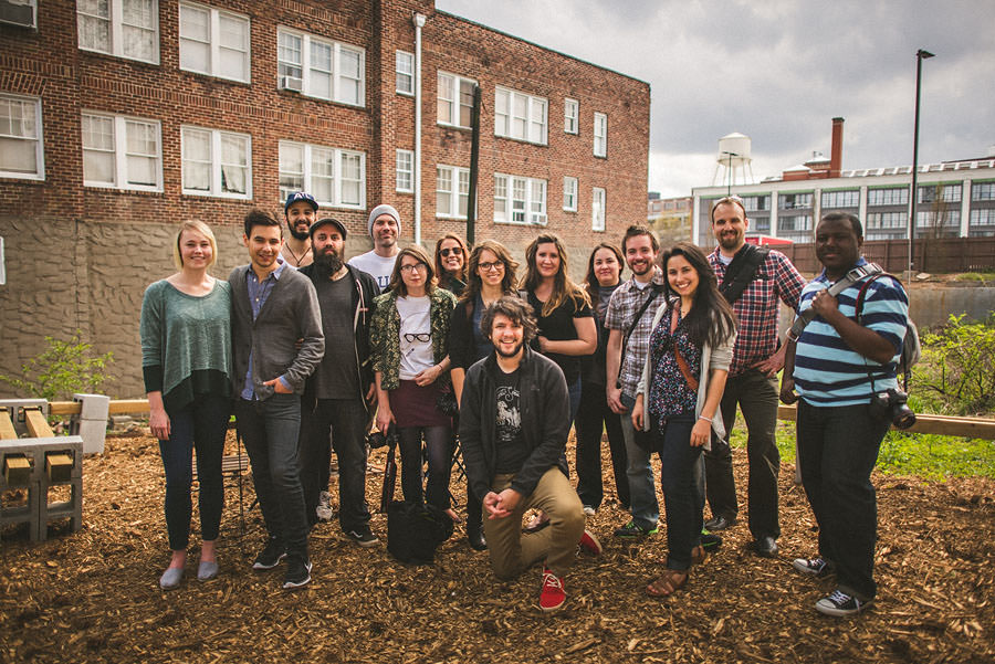 A group of 16 people standing and posing for a photo outdoors in front of a brick building on a cloudy day.