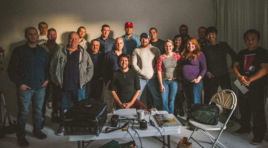 A group of 18 people posing for a photo in a well-lit room. Some sit and others stand, smiling at the camera. Various equipment and bags are visible in the foreground.