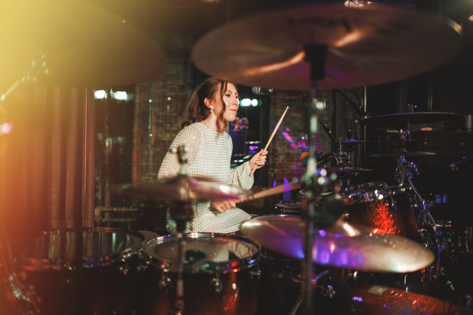 A woman in a light sweater plays a drum set on stage, holding drumsticks and smiling, with stage lights and drum equipment visible around her.