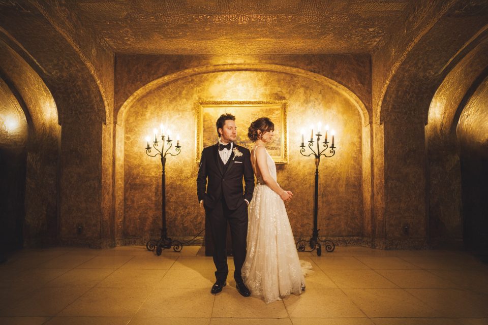 A couple in formal wedding attire stands back-to-back in an ornate room with gold walls, arched ceilings, candelabras, and a framed painting in the background.