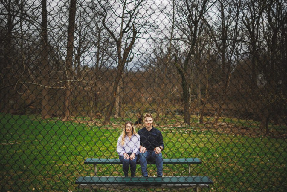 Two people sit side by side on a bench in a park, behind a chain-link fence with leafless trees and green grass in the background.