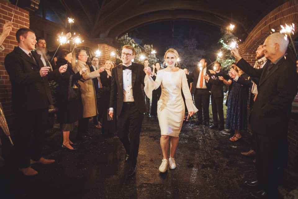 A newlywed couple walks under an archway at night as guests holding sparklers line both sides, celebrating their wedding.