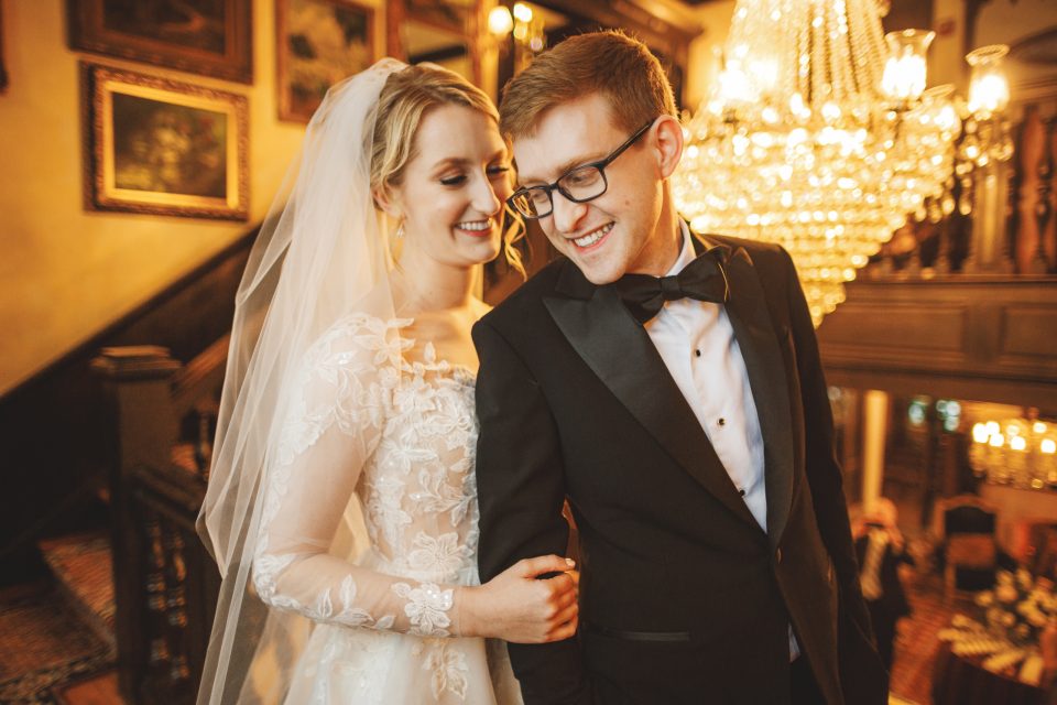 A bride in a lace wedding dress and veil stands arm in arm with a groom in a tuxedo, both smiling, in an ornate room with a chandelier and framed paintings.