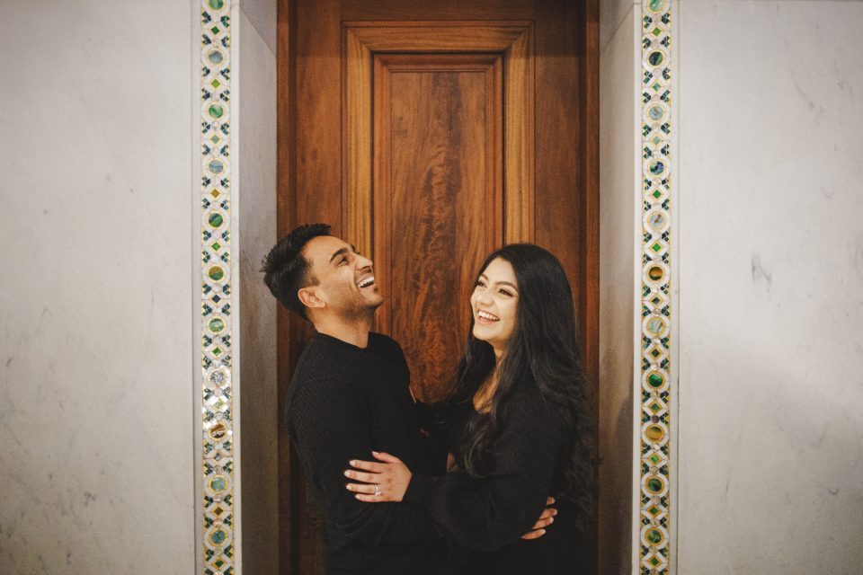 A man and a woman stand close together, smiling and laughing in front of a wooden door framed by decorative tiles.