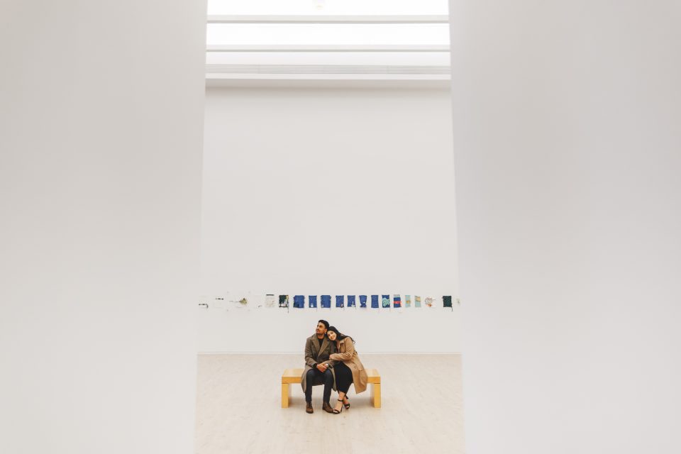 Two people sit closely together on a wooden bench in a spacious, minimalist art gallery with artworks displayed on the far white wall.