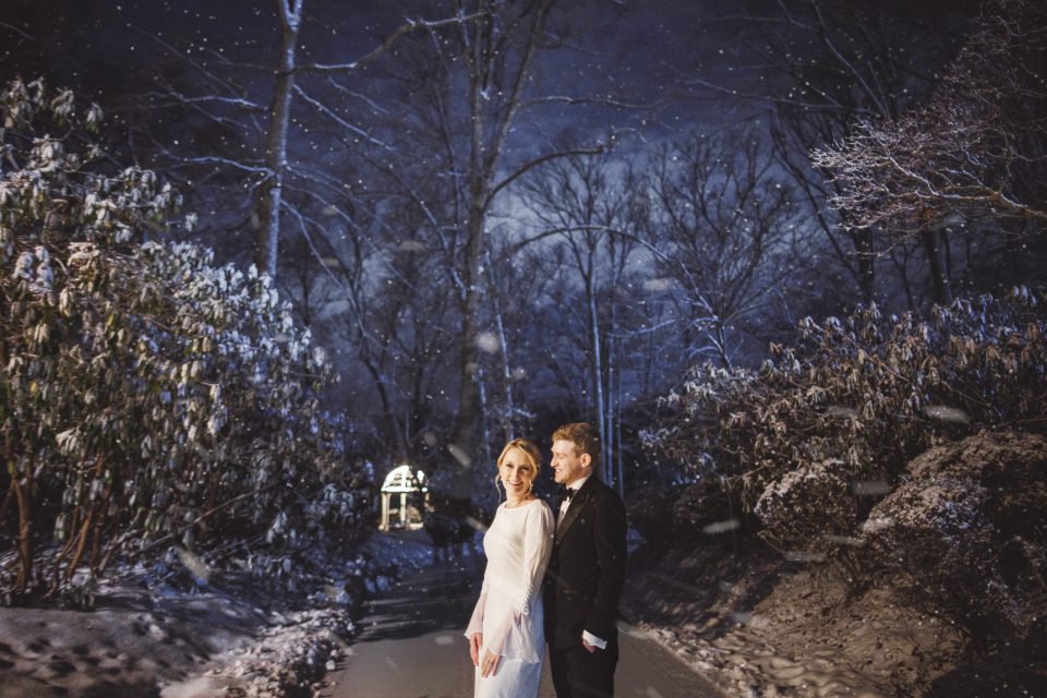 A bride and groom stand together on a snowy path at night, surrounded by snow-covered trees and falling snowflakes.