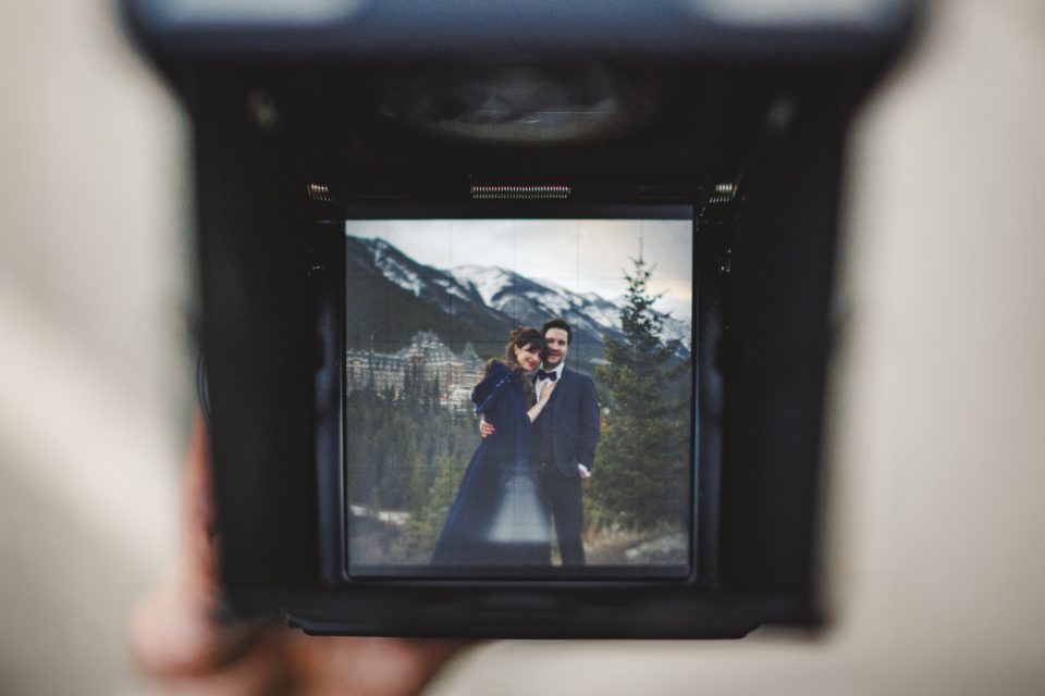 A couple poses in formal attire outdoors, with mountains and trees in the background, as seen through a camera viewfinder.