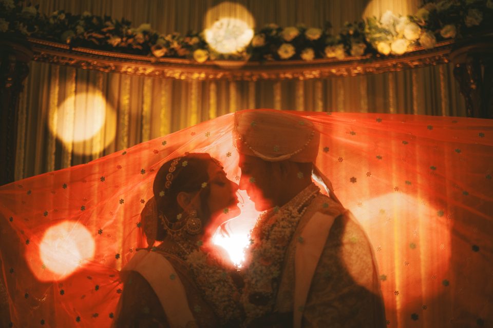 Two people in traditional wedding attire stand close under a veil, facing each other, with warm lighting and floral decorations in the background.
