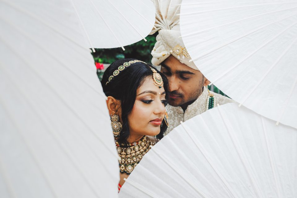 A bride and groom in traditional attire are framed by white umbrellas, standing close together and gazing softly downward.