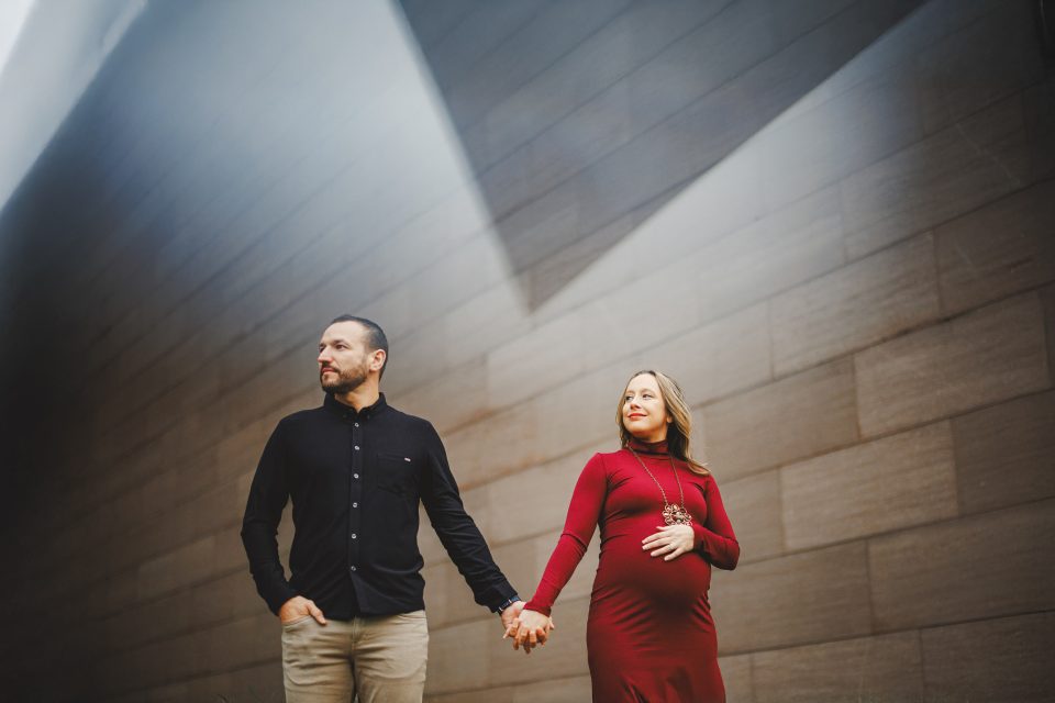 A man and a pregnant woman in a red dress hold hands while standing under angled light against a modern, gray stone wall.