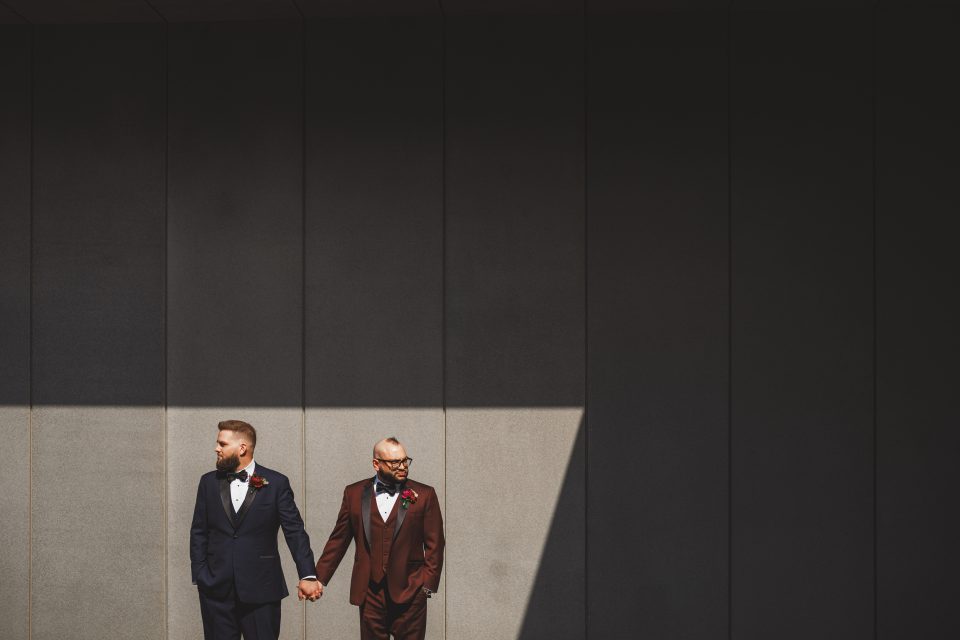 Two men in suits, one navy and one brown, stand holding hands against a tall, gray, shadowed wall with a beam of sunlight partially illuminating them.