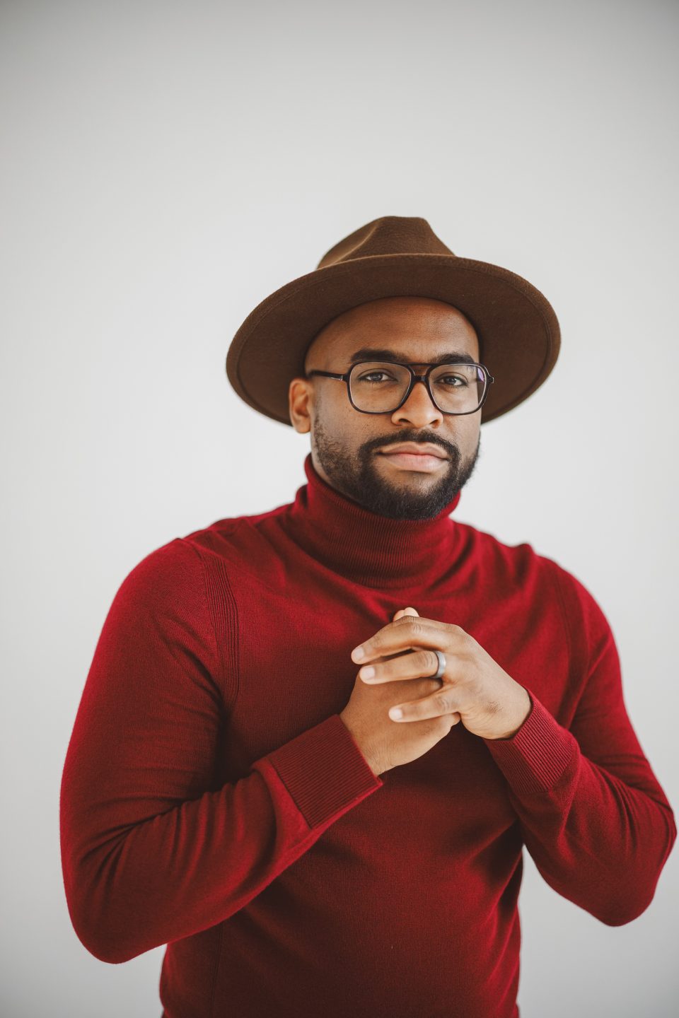 A man wearing glasses, a brown hat, and a red turtleneck sweater stands against a plain light background with his hands clasped together.