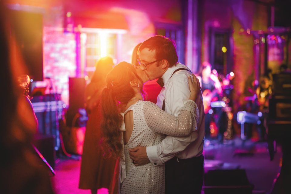 A couple dressed in formal attire share a kiss while dancing at a party or wedding reception with colorful lighting and a live band in the background.