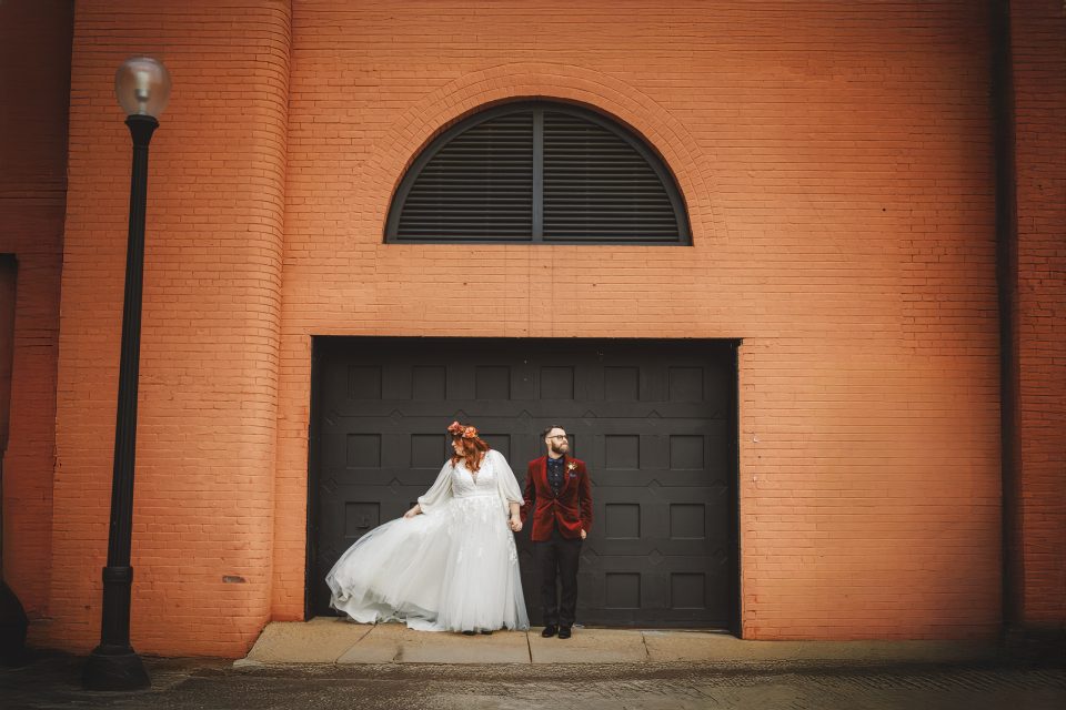 A bride in a white dress and a groom in a red suit stand side by side in front of a large black garage door set in an orange brick building.