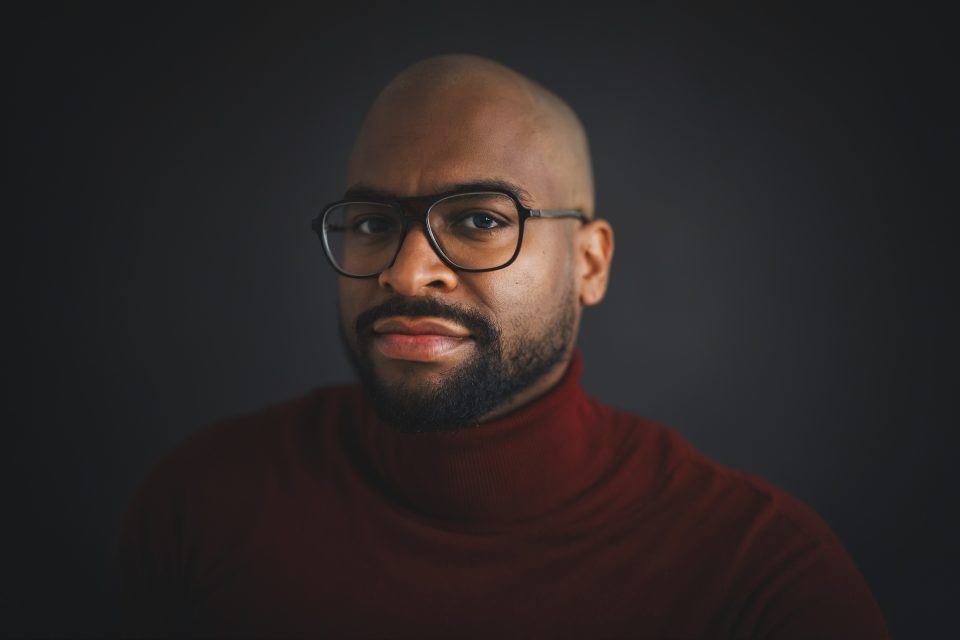A man wearing glasses and a dark red turtleneck poses against a dark background, looking directly at the camera with a neutral expression.