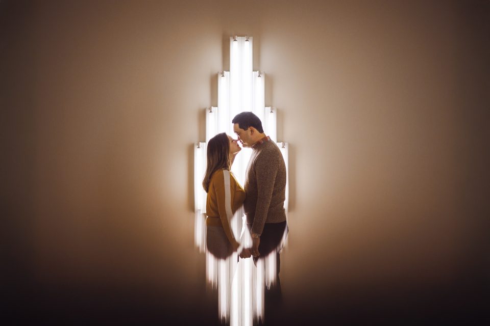 A couple stands close together in front of a wall-mounted vertical light fixture, holding hands and facing each other. The lights create a radiant, symmetrical backdrop.