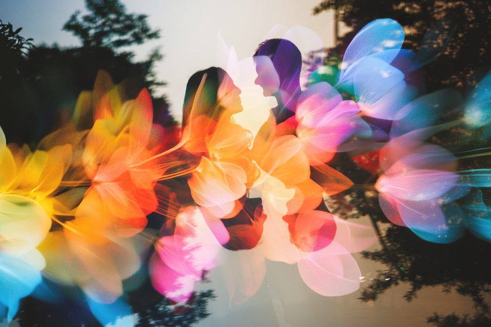 A couple faces each other and holds hands, surrounded by a vibrant, colorful overlay of abstract floral or light shapes against a dark outdoor background.