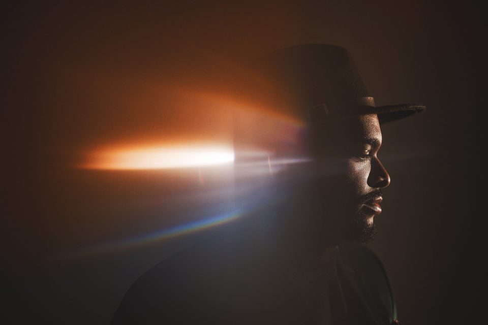 A man in profile view wearing a dark hat stands against a dark background, illuminated by a beam of orange and white light across the image.